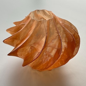 Könnte beinhalten: Ein orangefarbener, halbtransparenter Lampenschirm mit Spiraldesign. Der Lampenschirm hat eine Reihe von gebogenen, blütenblattartigen Abschnitten, die einen geschichteten Effekt erzeugen. Das Material scheint ein dünner, zarter Stoff zu sein, und die Gesamtform erinnert an eine Blume.