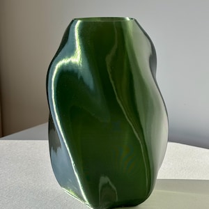 May include: A glossy, green vase with an organic, flowing shape. The vase has a wide, open top and a smooth, reflective surface. The vase is approximately 10 inches tall and is displayed on a white surface.