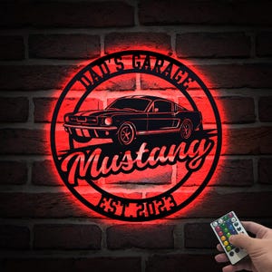 Peut inclure: Panneau métallique noir avec éclairage LED rouge, représentant une silhouette de voiture Mustang classique. Le panneau indique "Dad's Garage Mustang EST. 2023" dans un design circulaire, monté sur un mur de briques.