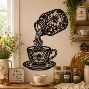 Könnte beinhalten: Ein Wandkunstobjekt aus schwarzem Metall, das ein geneigtes Glas zeigt, aus dem Flüssigkeit in eine Teetasse fließt. Sowohl das Glas als auch die Tasse sind mit filigranen Blumenmustern und Bienenillustrationen verziert.