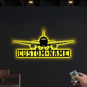 以下が含まれることがあります： 黄色いLEDライトで照らされた黒い金属製の飛行機のシルエット。「CUSTOM NAME」の文字が長方形のフレームに入っています。リモコンが右下に表示されています。