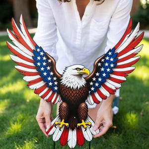 Puede incluir: Un adorno de jardín patriótico con un águila calva americana con las alas extendidas. Las alas presentan rayas rojas y blancas con campos de estrellas azules. El águila tiene la cabeza blanca, el cuerpo marrón y el pico y las garras amarillas. Se sujeta con una estaca de metal verde.