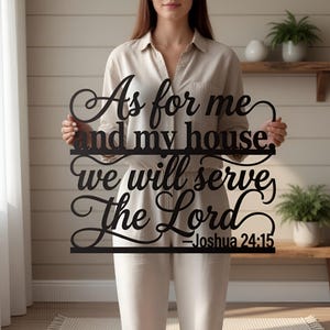 Peut inclure: Décoration murale en métal noir avec la citation "As for me and my house, we will serve The Lord - Joshua 24:15" en écriture élégante. Le panneau est tenu par une personne vêtue d'une tenue claire, sur un fond neutre.
