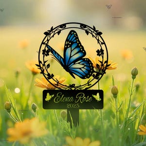 Puede incluir: Una estaca de jardín de metal negro con una silueta de mariposa y un letrero que dice "Elena Rose 2025". La estaca está rodeada de flores amarillas y hierba verde.
