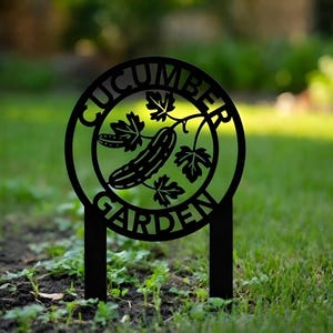 May include: Black metal garden sign with the words "CUCUMBER GARDEN" in a circular design. The sign features a cucumber and leaves illustration. The sign is staked into the ground, ideal for marking a vegetable patch.