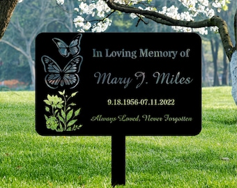 May include: A black memorial garden stake with rounded corners. The sign features butterfly and floral designs, along with the text "In Loving Memory of Mary J. Miles" and dates. The phrase "Always Loved, Never Forgotten" is also included.