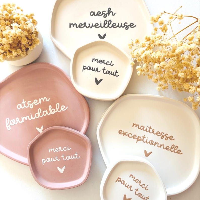 May include: Four decorative ceramic trays with different phrases in French. The trays are in shades of pink and white. The phrases are "aesh merveilleuse", "atsem formidable", "maitresse exceptionnelle", and "merci pour tout".