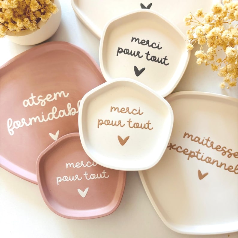 May include: Four ceramic trays with different messages written on them. The trays are in various shades of pink and white. The messages on the trays are "atsem formidabl", "merci pour tout", "merci pour tout", and "maitresse exceptionnelle".