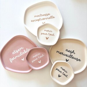 May include: Four different-sized, irregularly shaped ceramic trays with white text on a white background. The largest tray says "maitresse exceptionnelle" and the smallest tray says "merci pour tout". The other two trays say "atsem formidable" and "aesh merveilleuse".
