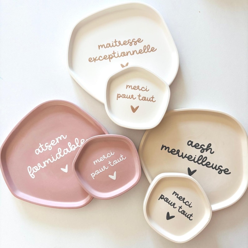 May include: Four different-sized, irregularly shaped ceramic trays with white text on a white background. The largest tray says "maitresse exceptionnelle" and the smallest tray says "merci pour tout". The other two trays say "atsem formidable" and "aesh merveilleuse".