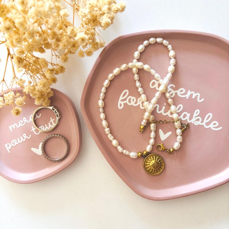 May include: Two pink ceramic dishes with white lettering. One dish has a pearl necklace with a gold sun pendant and the text "a sem formidable". The other dish has two rings and the text "merci pour tout".