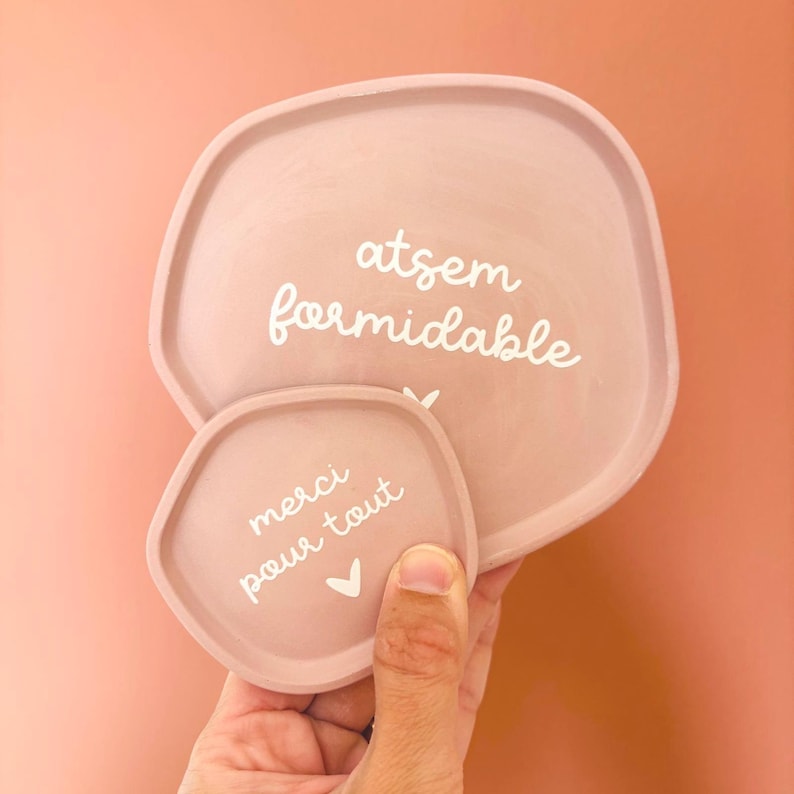 May include: Two pink ceramic trays with rounded edges. The larger tray has the text "atsem formidable" written in white. The smaller tray has the text "merci pour tout" written in white with a small heart symbol.