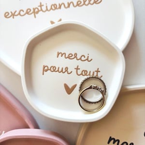 May include: White ceramic ring dish with a gold inscription that reads "merci pour tout" and a small gold heart. Two gold rings are sitting in the dish.