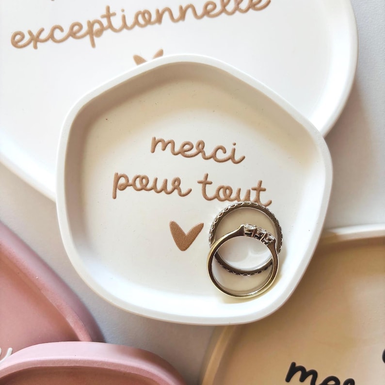 May include: White ceramic ring dish with a gold inscription that reads "merci pour tout" and a small gold heart. Two gold rings are sitting in the dish.