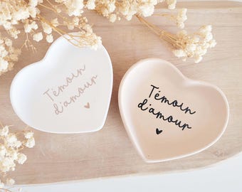 Może przedstawiać: Dwa sercowate naczynia na drobiazgi, jedno białe i jedno w kolorze różu, z francuskim tekstem "Témoin d'amour" i małym sercem. Naczynia znajdują się na jasnej drewnianej tacy, z delikatnymi białymi kwiatami w tle.