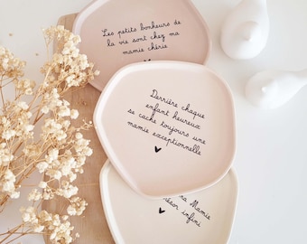 Puede incluir: Tres platos de cerámica color rubor con escritura negra. Los platos tienen bordes irregulares y presentan texto en francés. Dos figuras de pájaros blancos están en el fondo. Flores secas sobre una tabla de madera.
