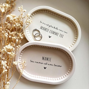 Peut inclure: Deux plateaux ovales blancs cassés avec des bords perlés. L'un porte l'inscription "la vie est plus belle avec une MAMIE COMME TOI" et un cœur. L'autre porte l'inscription "MAMIE ton amour est mon trésor" et un cœur. Deux bagues sont posées sur le plateau supérieur.