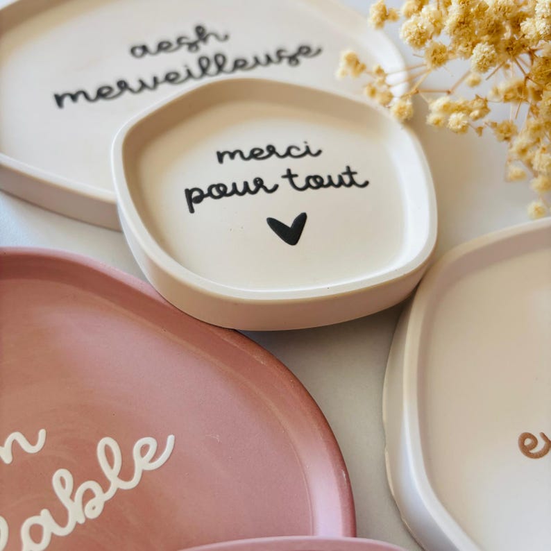 May include: Three small, white ceramic dishes with rounded edges. The dishes have black lettering. One dish says "aesh merveilleuse", another says "merci pour tout" with a small heart, and the third says "remarkable".