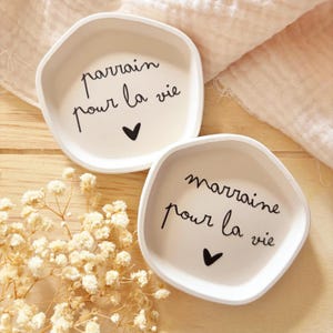 Peut inclure: Deux petits plats en céramique blanc cassé de forme irrégulière. L'un porte l'inscription "parrain pour la vie" et un cœur noir. L'autre porte l'inscription "marraine pour la vie" et un cœur noir. Sur une surface en bois.