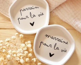 Może przedstawiać: Dwa kremowe, nieregularne ceramiczne naczynia. Jedno naczynie ma napis "parrain pour la vie" i czarne serce. Drugie naczynie ma napis "marraine pour la vie" i czarne serce. Naczynia są na drewnianej powierzchni.