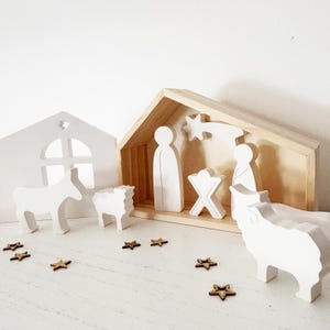 Peut inclure: Une crèche en bois avec des figurines blanches représentant Marie, Joseph, l'enfant Jésus, des animaux et une étoile. Les figurines sont placées dans une étable en bois et une maison blanche. De petites étoiles en bois sont éparpillées.