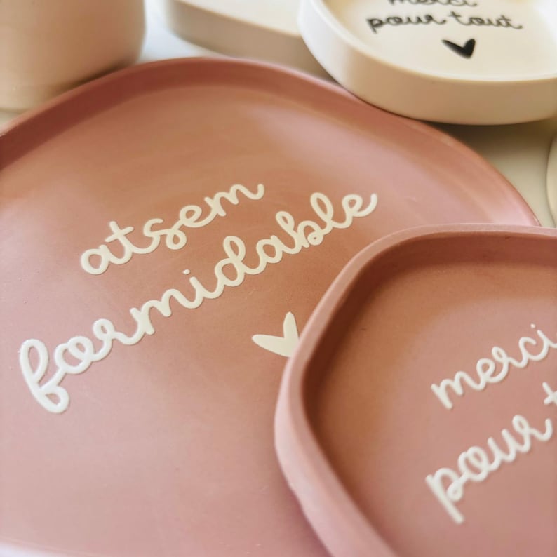 May include: Two pink ceramic trays with white lettering. The larger tray says "atsem formidable" and the smaller tray says "merci pour tout".