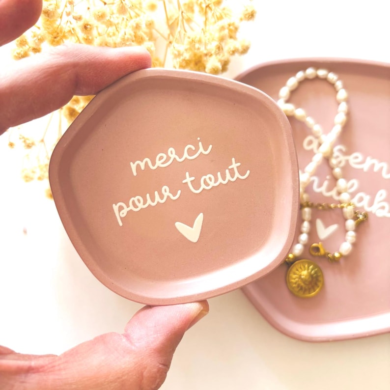 May include: A pink ceramic trinket dish with a white inscription that reads "merci pour tout" with a small white heart below the text.