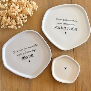 Op de afbeelding: Een set van vier witte keramische borden met onregelmatige randen. De borden hebben zwarte tekst in het Frans, waaronder zinnen als "MON PAPA" en "MON PAPA D'AMOUR", en een klein zwart hart. De borden zijn gerangschikt op een houten oppervlak.