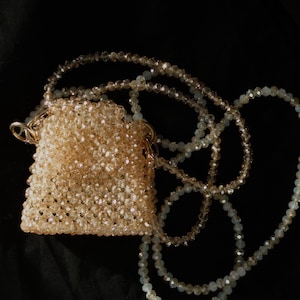 Può includere: Una piccola borsa a tracolla con perline e una catena dorata. La borsa è realizzata con perline color marrone chiaro e la catena è in metallo dorato. La borsa è appoggiata su una superficie nera.