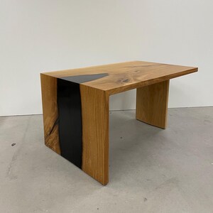 Könnte beinhalten: Ein Holz-Beistelltisch mit einer schwarzen Epoxidharz-Einlage. Der Tisch hat ein einzigartiges Design, bei dem die Beine seitlich aus der Tischplatte herausragen.