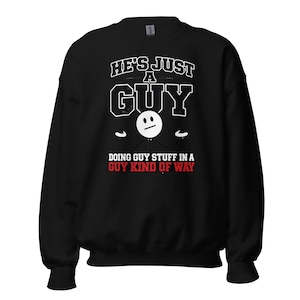 May include: A black long-sleeve sweatshirt featuring a white graphic of a stick figure with a shrug gesture. The text reads HE'S JUST A GUY in white and DOING GUY STUFF IN A GUY KIND OF WAY in red and white lettering on the front of the garment.