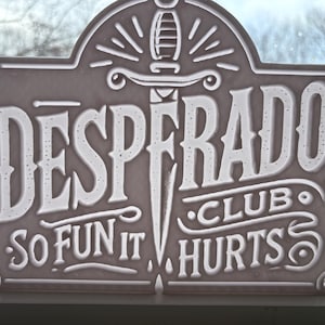 Może przedstawiać: Prostokątny znak z napisem "DESPERADO CLUB" i "SO FUN IT HURTS" w białych, stylizowanych literach. Sztylet jest wyśrodkowany na znaku. Znak jest w kolorze jasnoszarym.