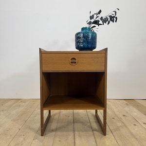 May include: A wooden nightstand featuring a single drawer with a circular recessed handle and an open storage shelf below. The nightstand has a light brown wood grain finish and is topped with a textured teal ceramic vase containing dark foliage.
