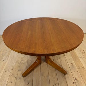 May include: A round wooden dining table with a warm brown finish and a visible wood grain pattern. The table features a central seam indicating an extendable design and is supported by a sturdy pedestal base with four angled legs.