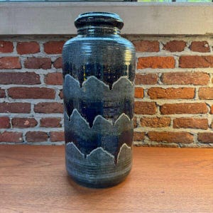 Può includere: Un vaso cilindrico in ceramica con smalto blu scuro e grigio antracite. La superficie presenta un motivo orizzontale ripetitivo di linee ondulate grigio chiaro che contrastano con la base scura, lucida e strutturata.
