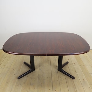 May include: A dark brown wooden dining table with a smooth, rounded rectangular top featuring a prominent wood grain pattern. The table is supported by two black, Y-shaped pedestal bases that extend outwards into angled feet at the bottom.
