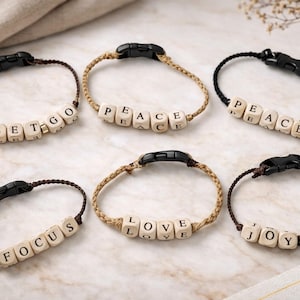 May include: Six handmade bracelets with black clasps and wooden letter beads spelling out "LET GO", "PEACE", "FOCUS", "LOVE", and "JOY". The bracelets are made with brown, tan, and black cords.