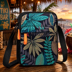 May include: A rectangular crossbody bag featuring a dark navy blue base with a repeating pattern of teal, light green, and tan tropical leaves. The bag includes a black adjustable strap, a front zippered pocket, and two bright orange zipper pulls.