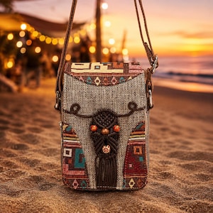 May include: A rectangular crossbody bag with a woven grey fabric body and a brown leather strap. The bag features colourful geometric patterns in red, orange, and teal. A decorative macrame element with beads adorns the front. The background is a beach scene at sunset.
