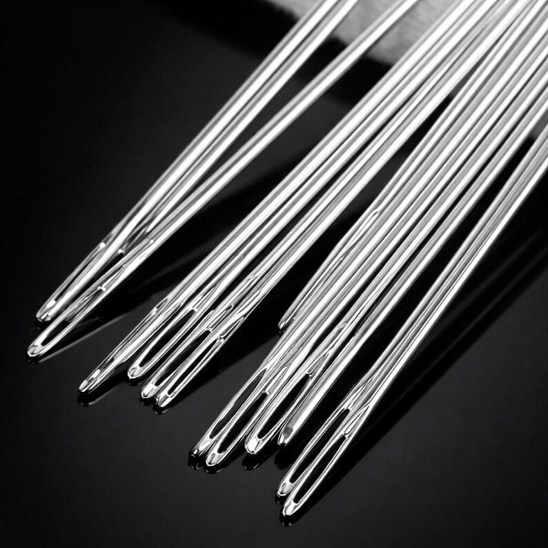 May include: A collection of long, silver sewing needles with large eyes, arranged on a dark, reflective surface. The needles are made of shiny metal and are slightly overlapping. The image highlights the needles' polished finish.