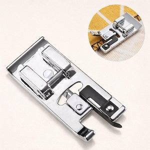 Pode incluir: Um calcador de máquina de costura prateado e brilhante com um detalhe preto. O calcador tem uma forma retangular com vários componentes metálicos e uma alavanca curva. Uma pequena inserção circular mostra um close-up do calcador.