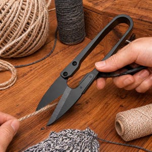 May include: Black craft scissors cutting twine on a wooden surface. The scissors have a curved handle and are next to spools of twine in various colors. The scene suggests a crafting or DIY project.