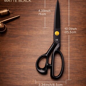 Puede incluir: Tijeras de sastre de acero al manganeso en negro mate con un tornillo de pivote amarillo. Las tijeras miden 25,5 cm de largo total, con una hoja de 11 cm y un ancho de 8,5 cm. El texto MANGANESE STEEL TAILOR y MATTE BLACK es visible.