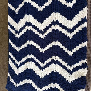 May include: A navy blue and white chevron-patterned blanket. The blanket is made of a thick, soft material, with a wavy edge. The design features alternating bands of navy blue and white, creating a zig-zag pattern across the surface.