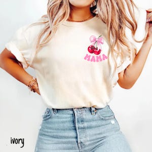 May include: Ivory-colored t-shirt featuring a graphic of two red cherries with a pink bow and the word "MAMA" in pink. The shirt has rolled-up sleeves and is paired with blue denim jeans. The word "ivory" is printed at the bottom left.