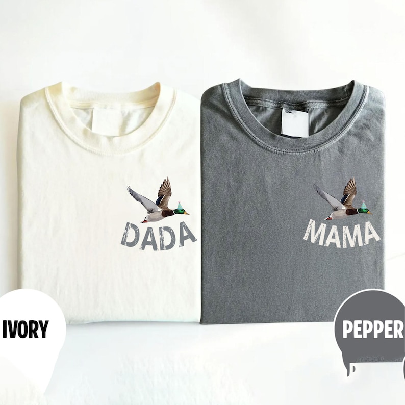 Puede incluir: Dos camisetas dobladas, una marfil y otra gris pimienta, cada una con un gr&aacute;fico de pato volando. La camiseta marfil tiene la palabra "DADA" impresa debajo, mientras que la gris dice "MAMA".