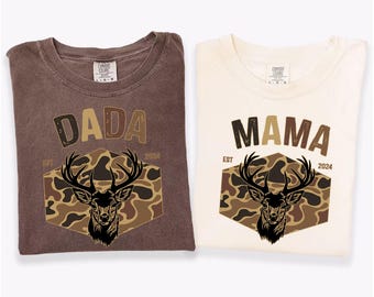 May include: Two t-shirts, one in espresso and one in ivory, featuring a deer head design. The espresso shirt displays "DADA", while the ivory shirt reads "MAMA". Both shirts include "EST 2024" below the text, creating a matching set.