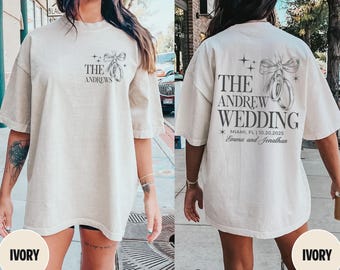 May include: Ivory-colored t-shirt featuring the text "THE ANDREWS" and a graphic of wedding rings and a bow. The back of the shirt displays "THE ANDREW WEDDING" with the date and location, and the names "Emma and Jonathan".