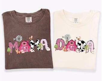 May include: Two t-shirts, one espresso and one ivory, with the words "MAMA" and "DADA" in a farm-themed design. The design includes a pink barn, windmill, and farm animals. The espresso shirt has a brown color, and the ivory shirt has a cream color.