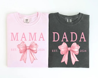 May include: Two t-shirts, one light pink and one dark gray, with the words "MAMA" and "DADA" in pink. Each shirt features a pink bow graphic and the text "EST. 2024". The bottom of the shirts read "blossom" and "pepper".